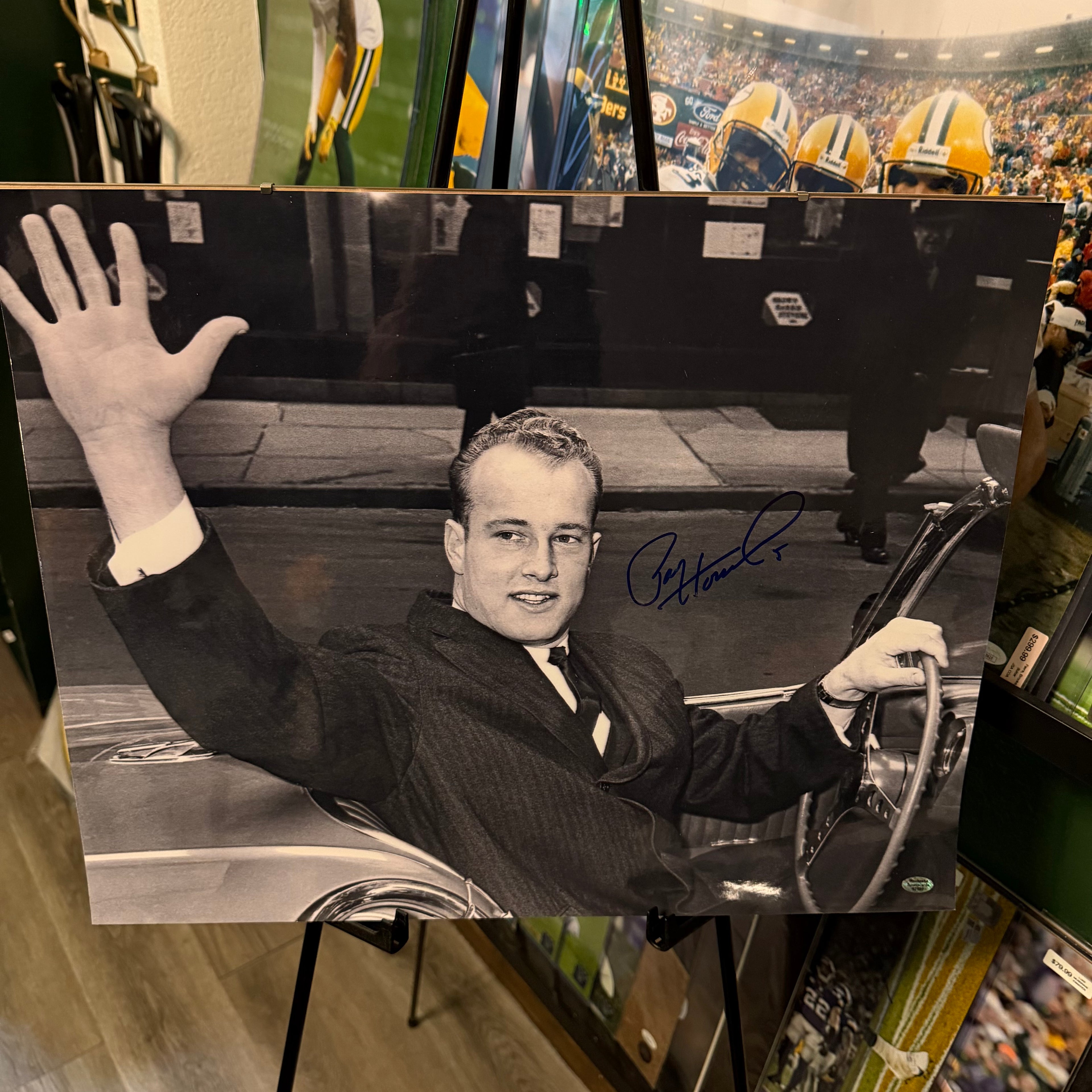 Autographed photo of Paul Hornung in a suit waving from a car, with sports memorabilia in the background.